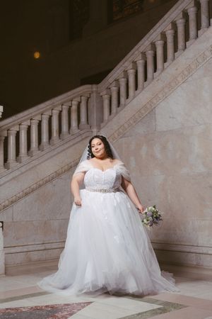 Plus-size bride in white off-shoulder ballgown holding bouquet on marble staircase.
