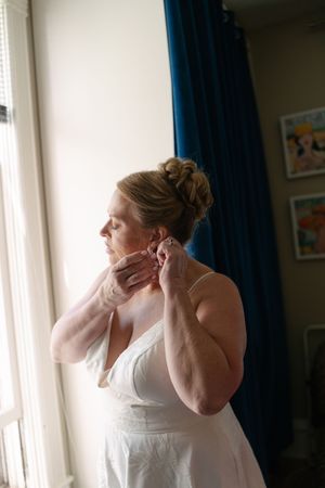 Bride in white gown putting on earrings by a bright window with blue curtains on her wedding day.