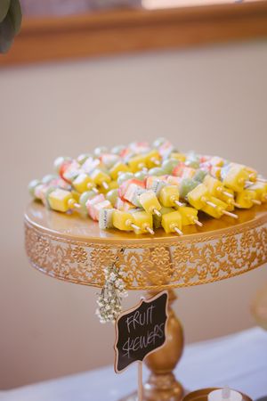 Colorful fresh fruit skewers displayed on decorative gold cake stand with chalkboard sign at wedding.