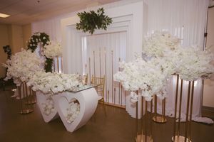 Elegant wedding venue with white floral arrangements on gold stands, heart-shaped decor, and draped white fabric backdrop.