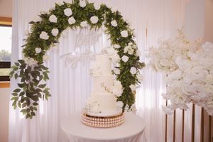 Elegant three-tier white wedding cake displayed under a lush floral arch with white roses and greenery on a decorative gold stand.