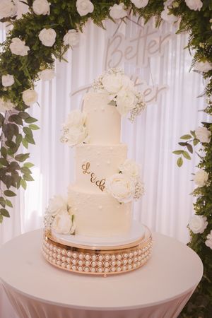 Four-tier white wedding cake decorated with roses and gold monogram initials on a pearl cake stand.