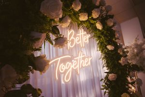 Neon sign reading Better Together surrounded by white roses and greenery at a romantic wedding reception.