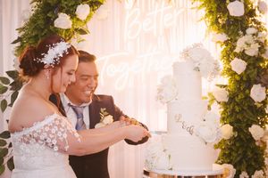 Bride and groom cutting white floral wedding cake under neon sign and greenery arch backdrop.