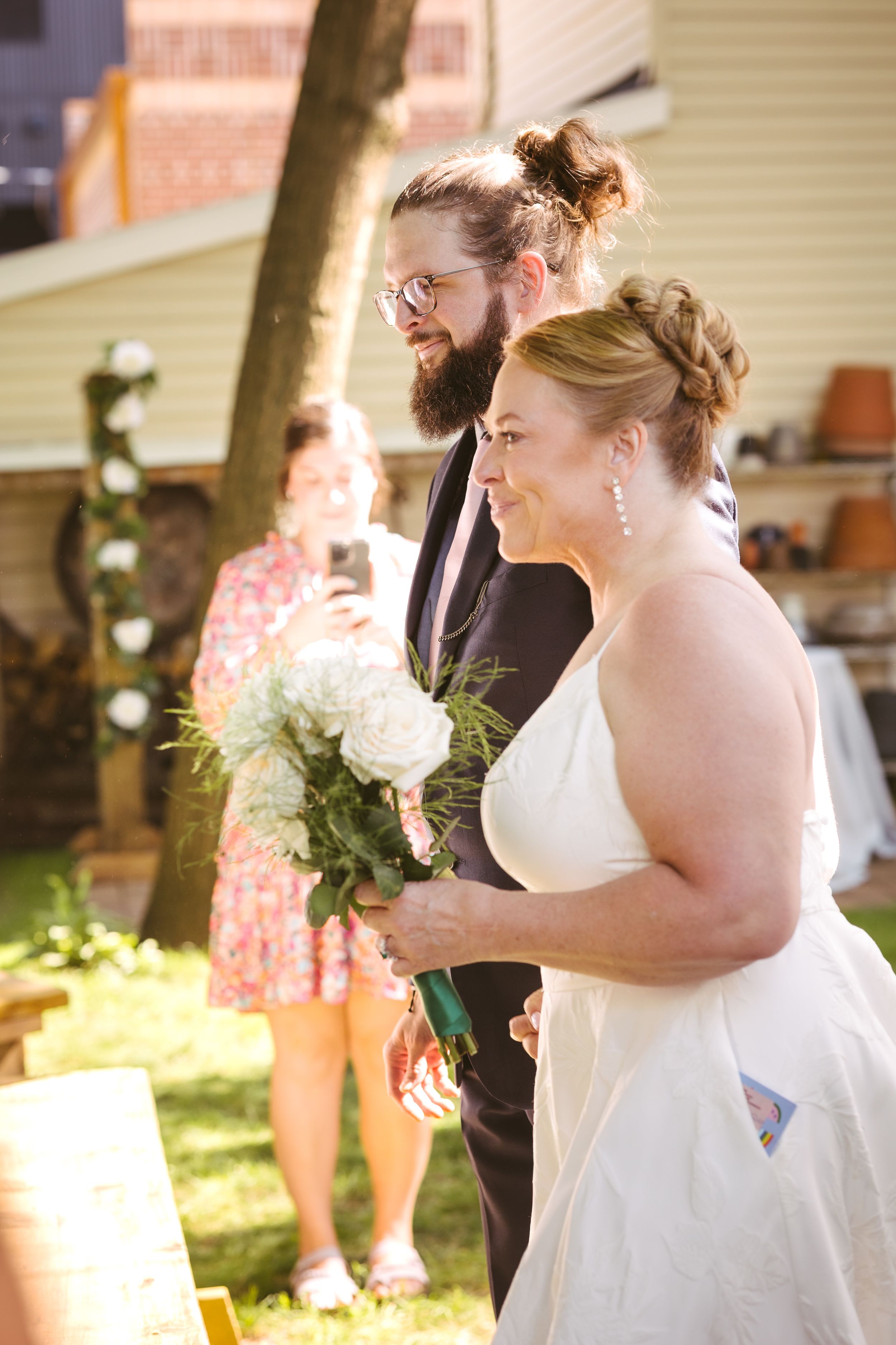 Smiling bride holding white bouquet walks with bearded groom in black suit at outdoor backyard wedding ceremony.