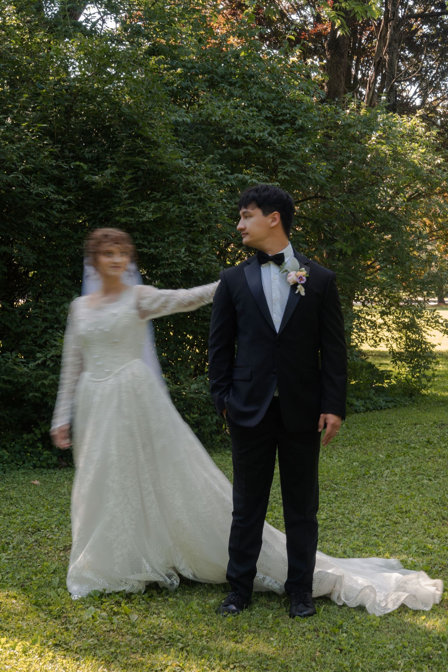 A bride in a white lace gown twirls beside a groom in a black tuxedo on a green lawn with trees behind them.
