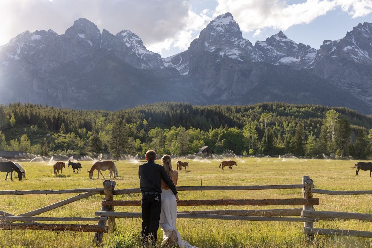 Couple embracing at a wooden fence overlooking horses grazing with the Grand Teton mountains in the background.