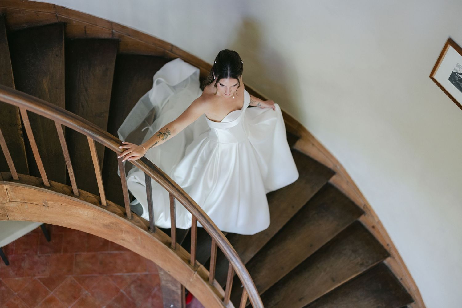 Bride in white wedding dress descending elegant spiral wooden staircase, holding gown, viewed from above.