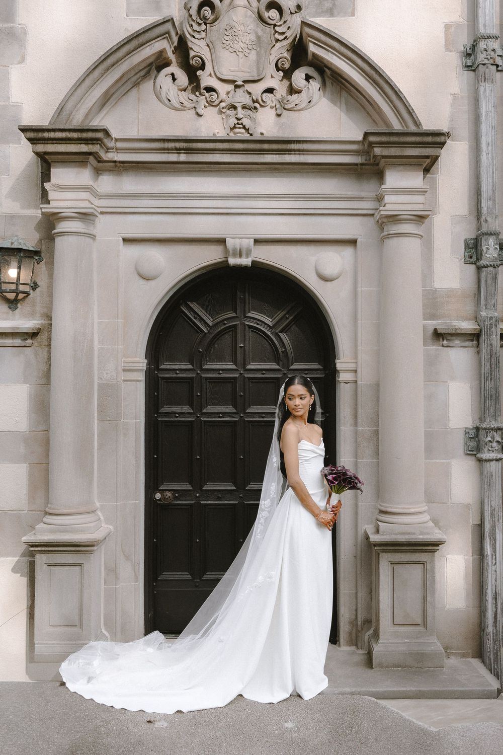 Bride in white gown and long veil holds bouquet before ornate stone archway with dark wooden door.
