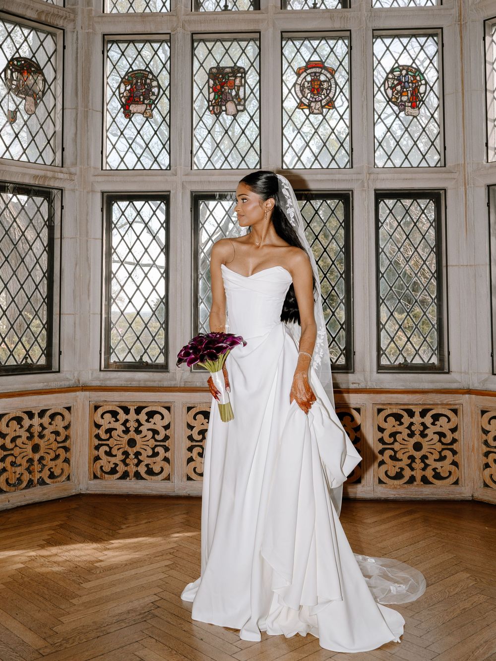 Bride in strapless white gown holding purple bouquet before stained glass windows in elegant venue.