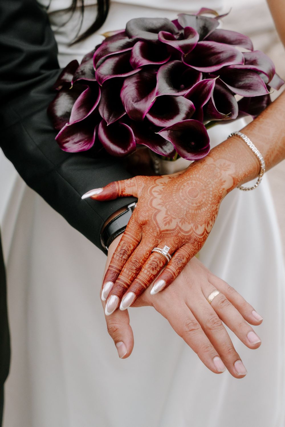 Bride's henna-adorned hands with wedding ring and white nails beside a dark purple calla lily bouquet.