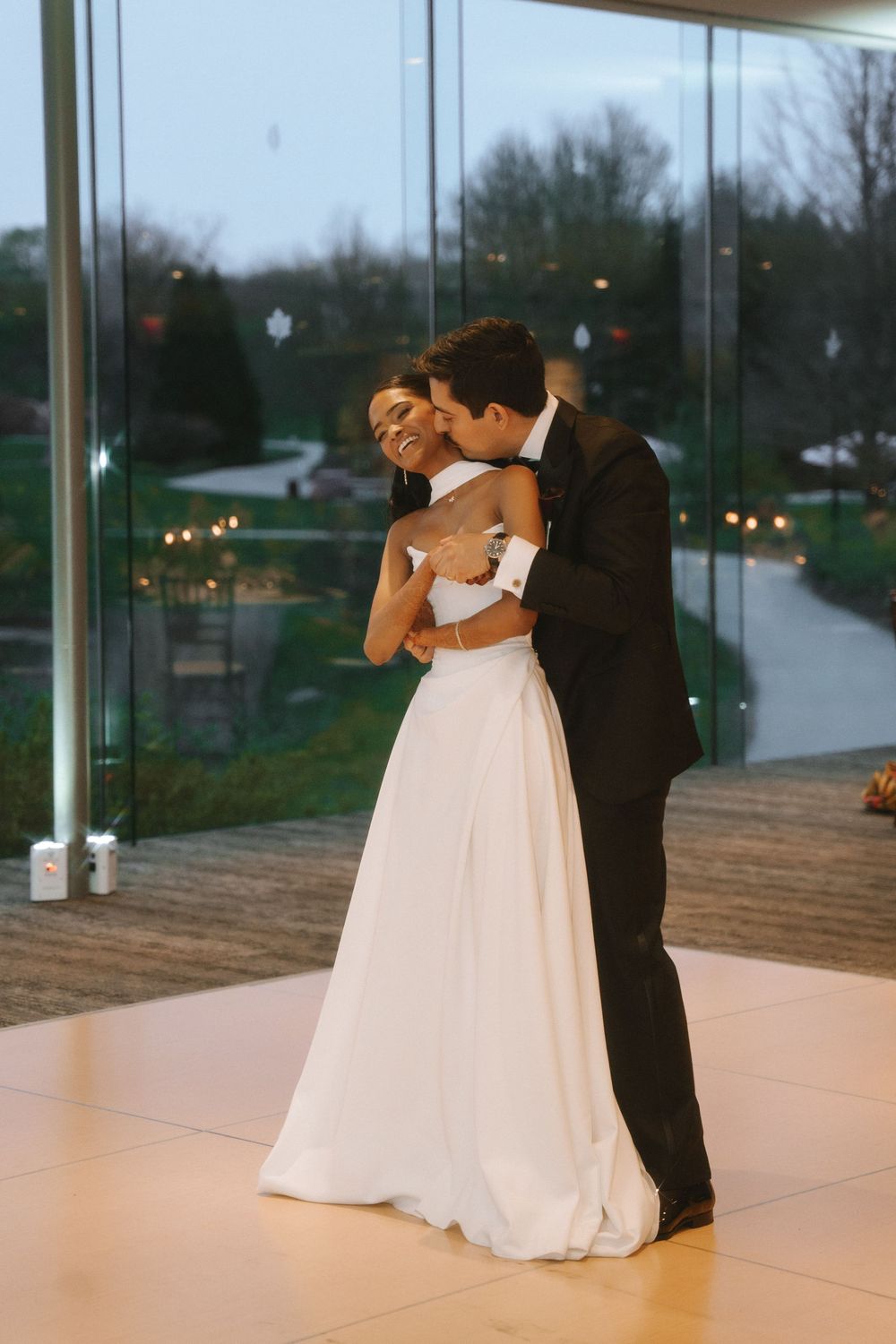 Bride and groom share first dance in elegant glass venue at dusk.