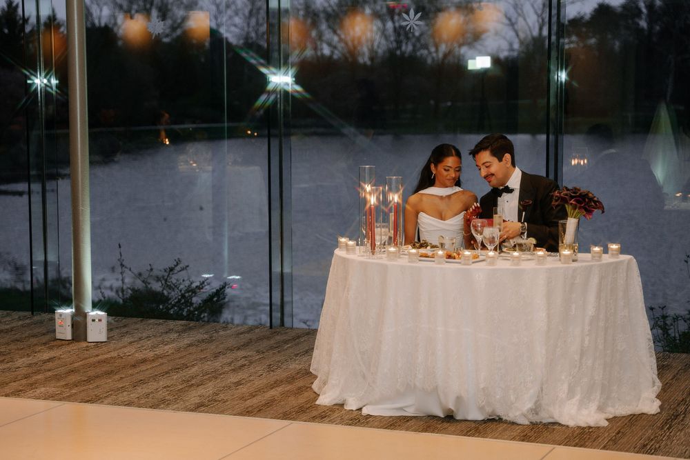 Bride and groom share an intimate moment at candlelit sweetheart table with white linen during evening wedding reception.