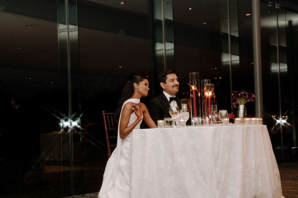 Bride and groom seated at sweetheart table during elegant wedding reception with candlelight and floral decor.