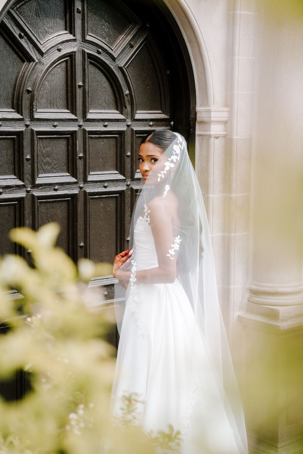 Bride in white gown and floral veil poses by ornate wooden church door.