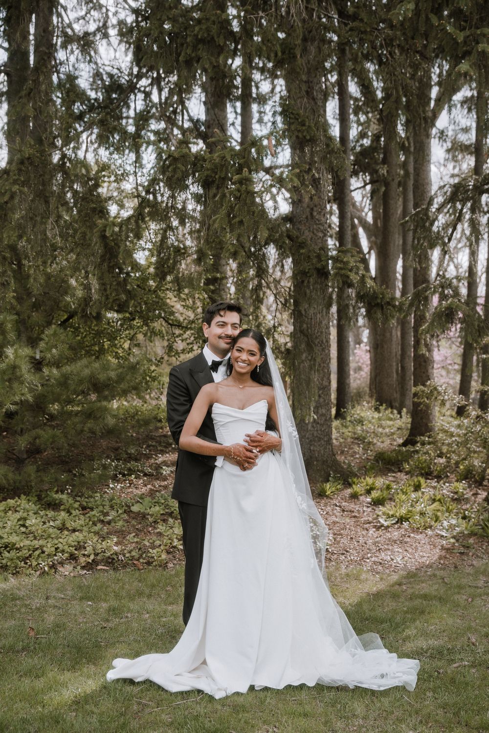 Newlywed couple embraces in lush forest setting, bride in strapless gown with veil.
