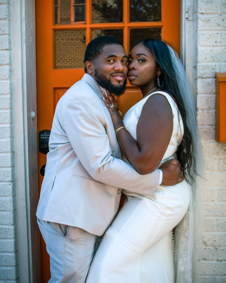 A couple in wedding attire embraces in front of an orange door, the bride in white and groom in a beige suit.