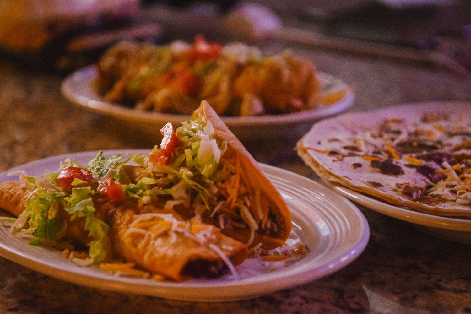 Colorful Mexican dishes with tacos and enchiladas topped with fresh lettuce and salsa on white plates.