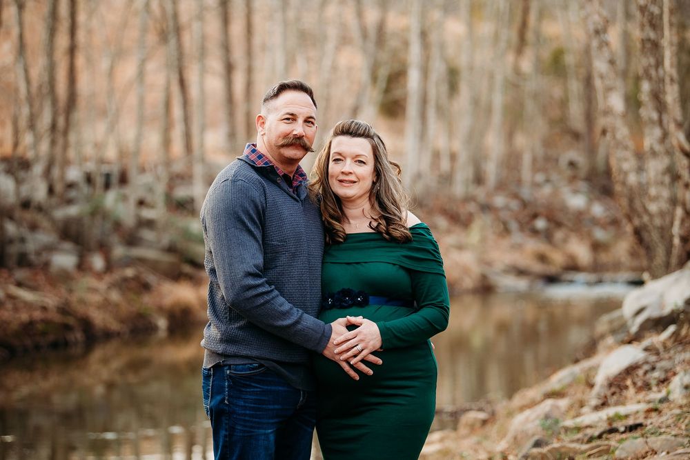 Couple embracing outdoors near a stream during a winter maternity photography session.