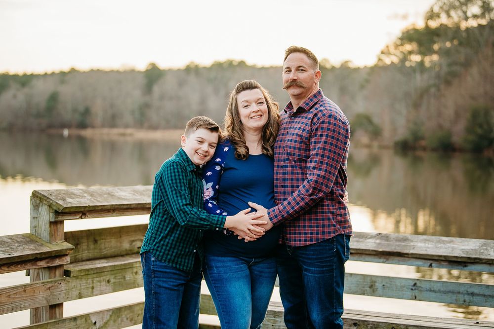 Pregnant woman with husband and son smiling together on a wooden dock by a lake at sunset.
