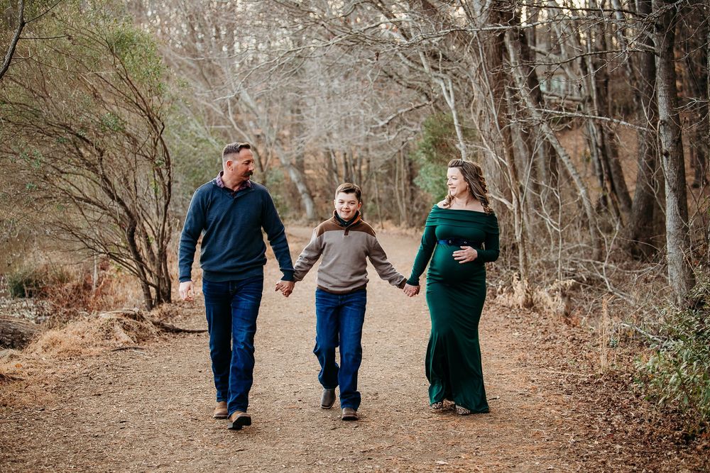 Family of three holding hands walking along a wooded trail during a maternity photo session.