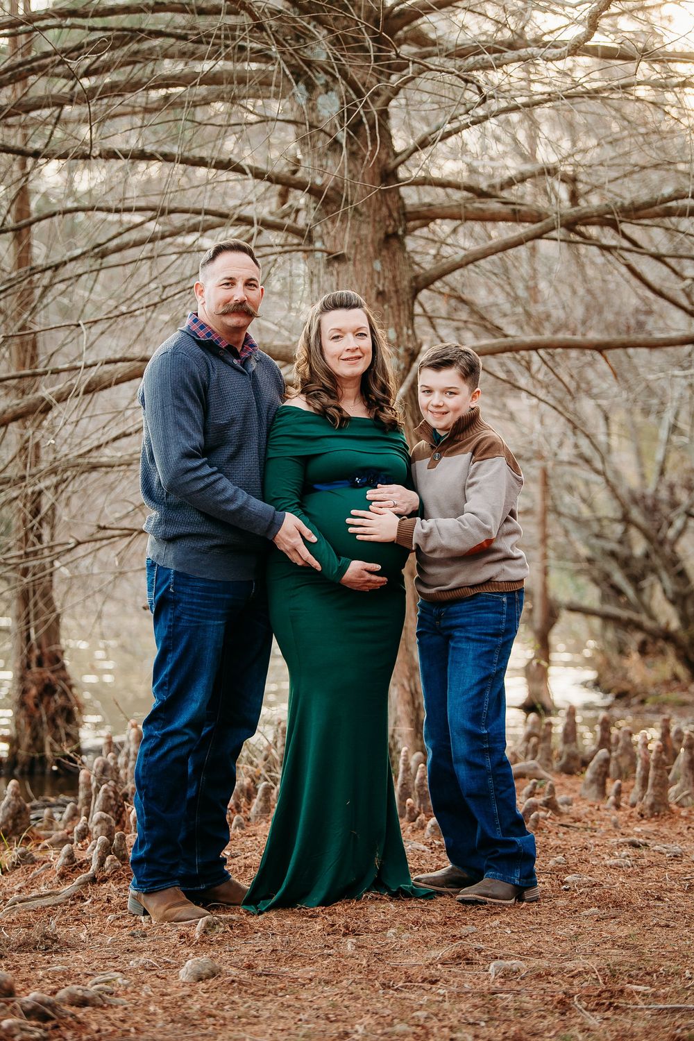 Pregnant woman in green gown with husband and son posing near trees for a maternity portrait.