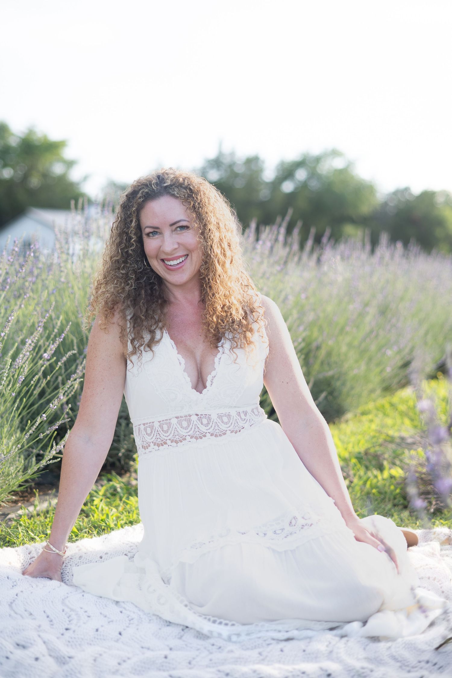 Smiling woman with curly hair in white lace dress sitting in lavender field on sunny day.