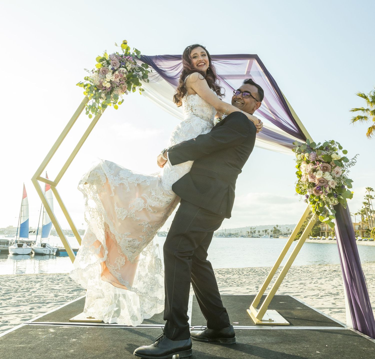 Bride and groom posing joyfully under a gold hexagonal arch with purple draping and flowers by the waterfront.