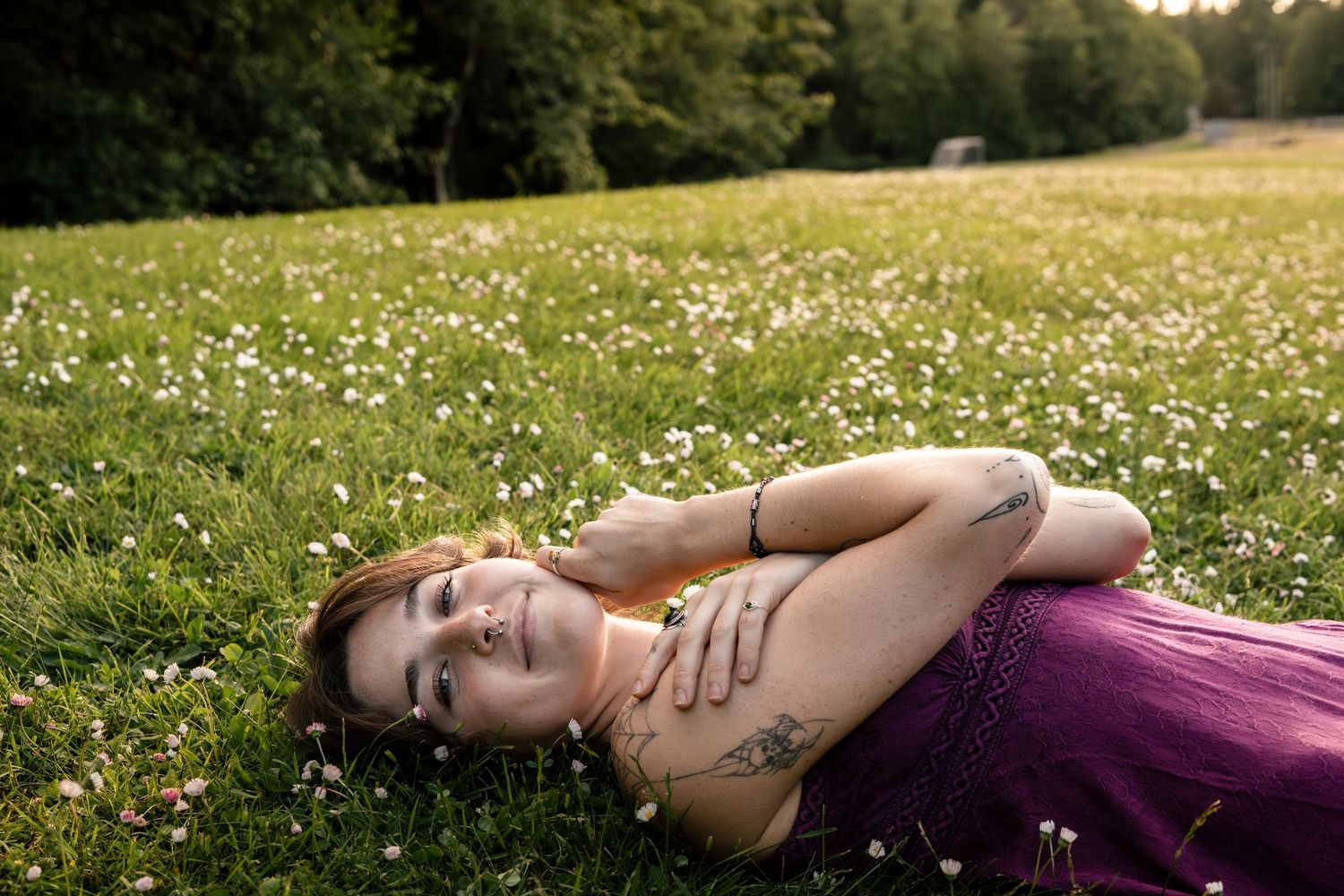Young woman with tattoos lies in a wildflower meadow wearing a purple dress at sunset.