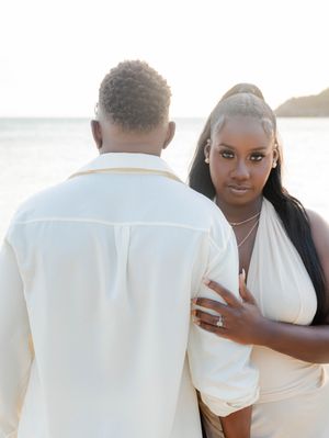 Couple in white outfits posing on a beach at sunset, woman with long hair and engagement ring looking back.