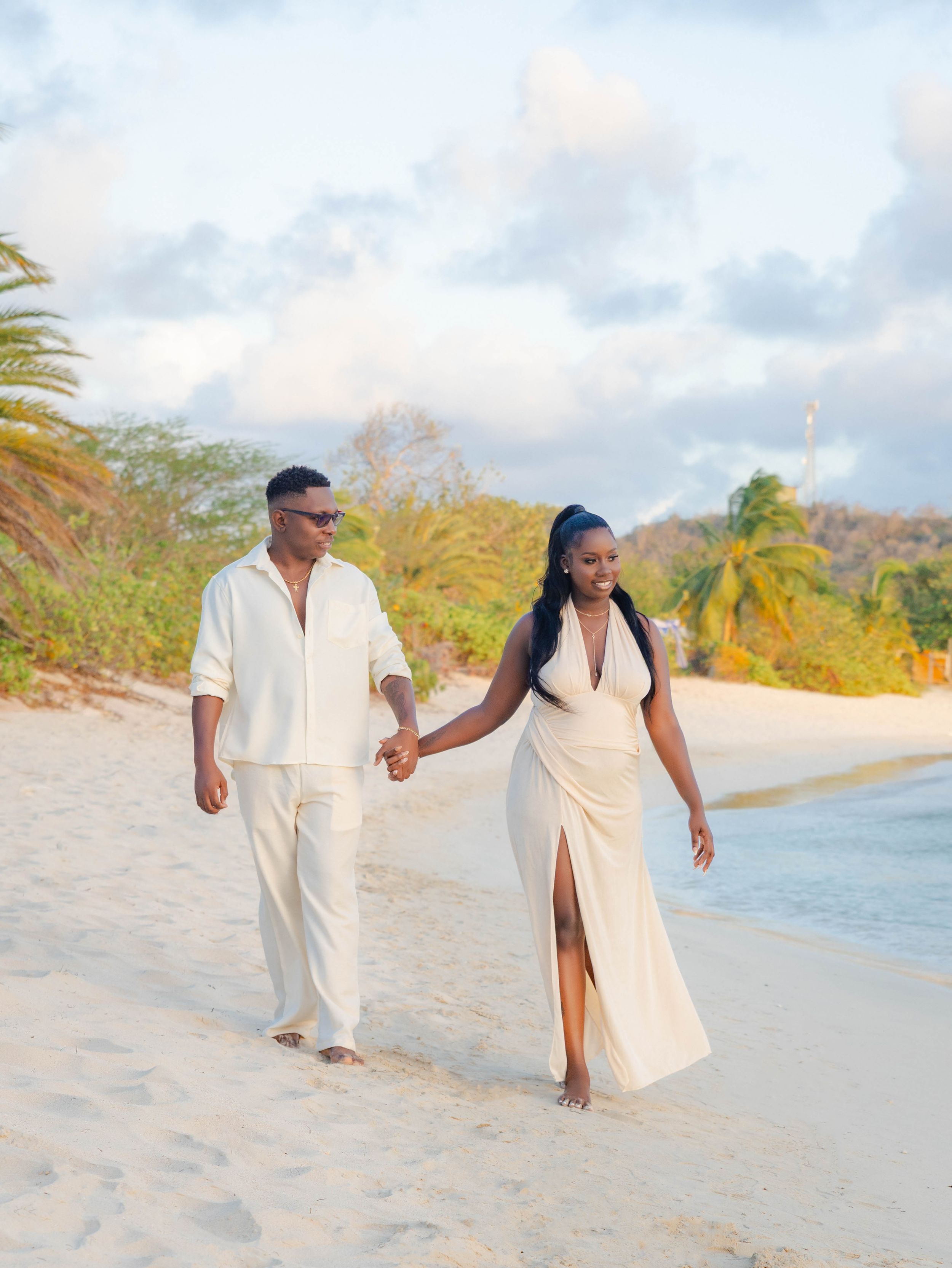 Couple in white outfits holding hands walking on a tropical beach at golden hour.