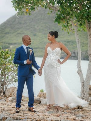 Couple in wedding attire holding hands outdoors by turquoise water, groom in blue suit, bride in white lace gown.
