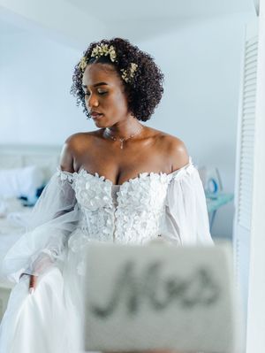 Bride in off-shoulder floral wedding gown with natural curly hair adorned with flowers, holding a Mrs. sign.