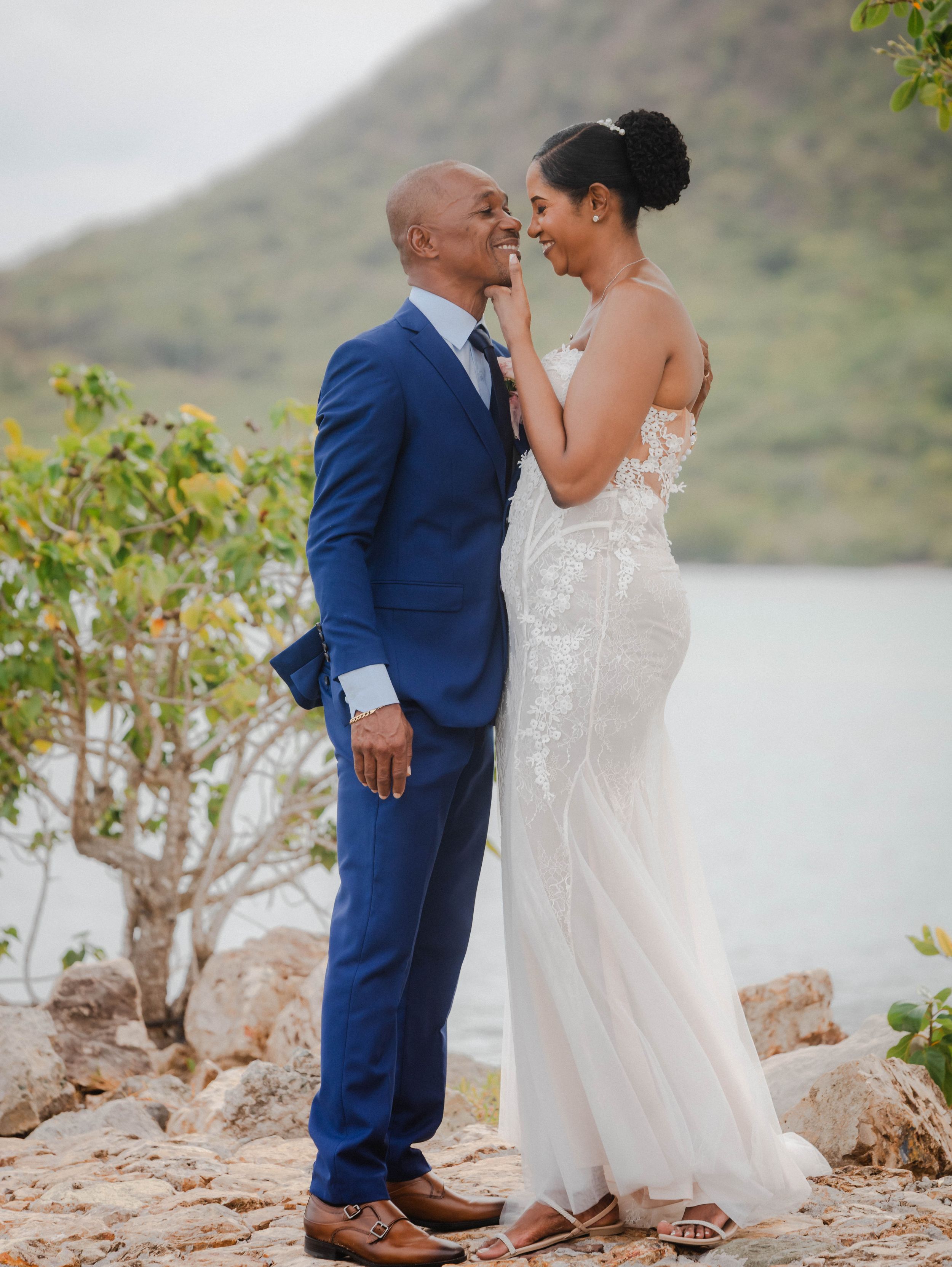 Couple in wedding attire smiling at each other on rocky shoreline with lush green hills and water in background.