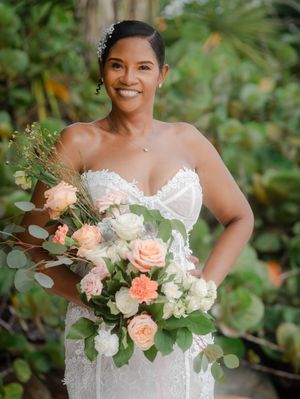 Smiling bride in strapless lace gown holding peach and white rose bouquet with greenery outdoors.
