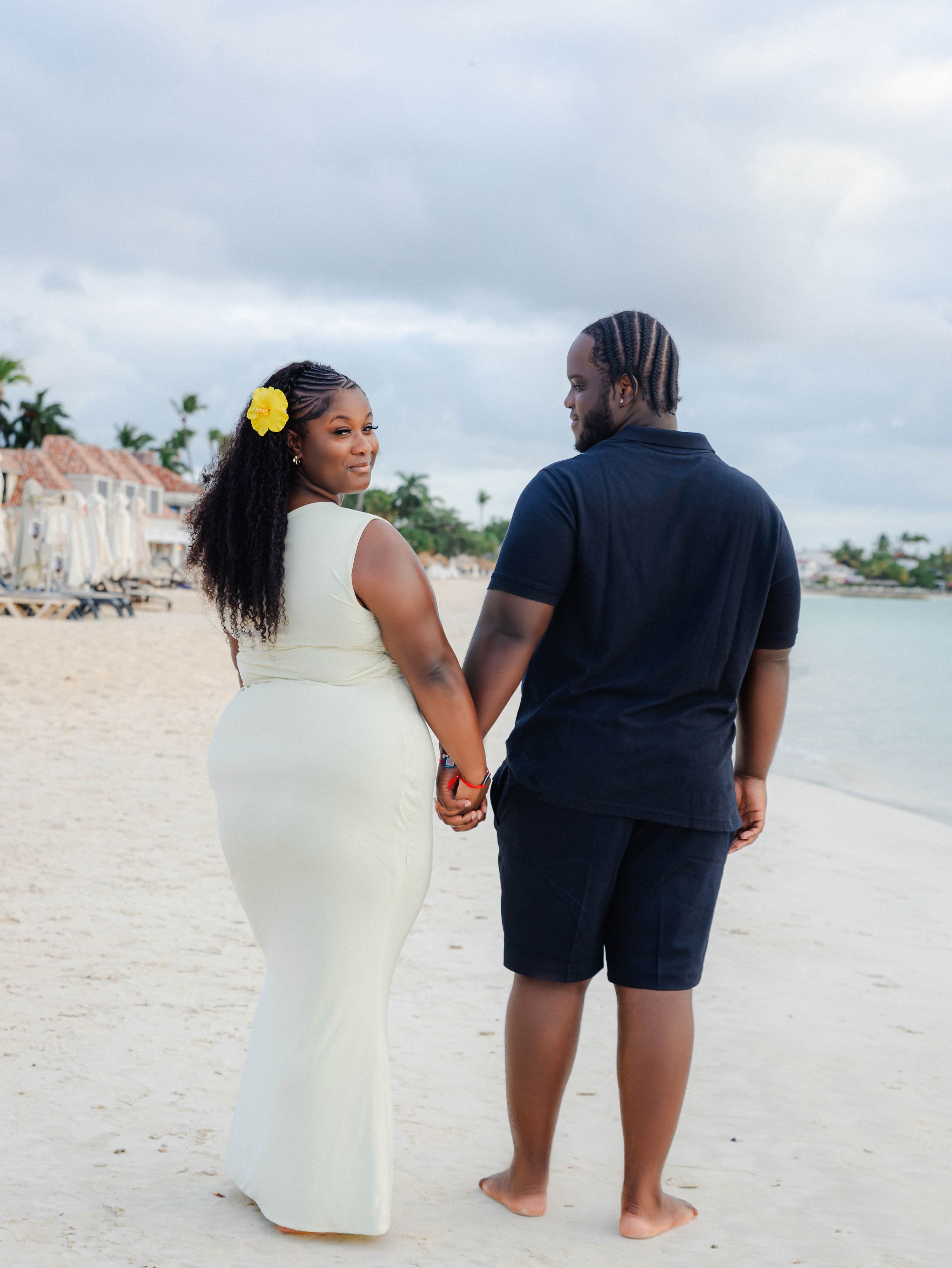 Couple holding hands on a sandy beach, woman in white dress with yellow flower, man in navy outfit, tropical resort backdrop.