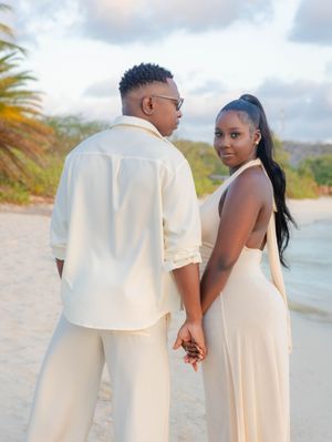 Couple in matching cream outfits holding hands on a tropical beach at sunset, woman in backless halter dress.