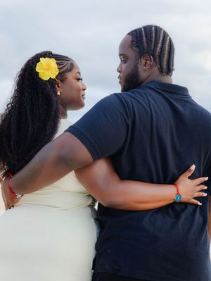 A couple embraces on a beach, the woman wearing a yellow hibiscus flower in her hair and a white dress.