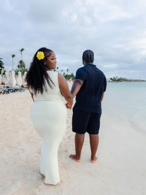 Couple holding hands on a tropical beach, woman in white dress with yellow flower in hair.