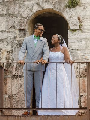 Newlywed couple posing on a balcony, groom in gray suit with green bowtie, bride in white lace wedding gown.