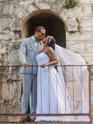 Bride and groom sharing a kiss on a balcony, groom in gray suit, bride in white lace gown with veil.