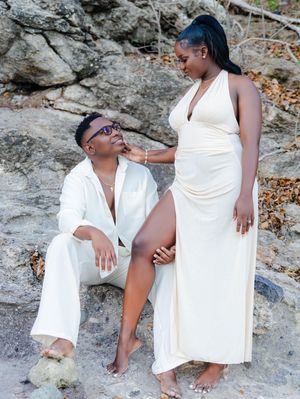 A couple in matching white outfits poses on rocky terrain, with the pregnant woman in a flowing gown.