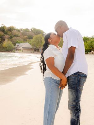 Couple kissing on a sandy beach while holding hands, wearing white tops and jeans with ocean waves in the background.