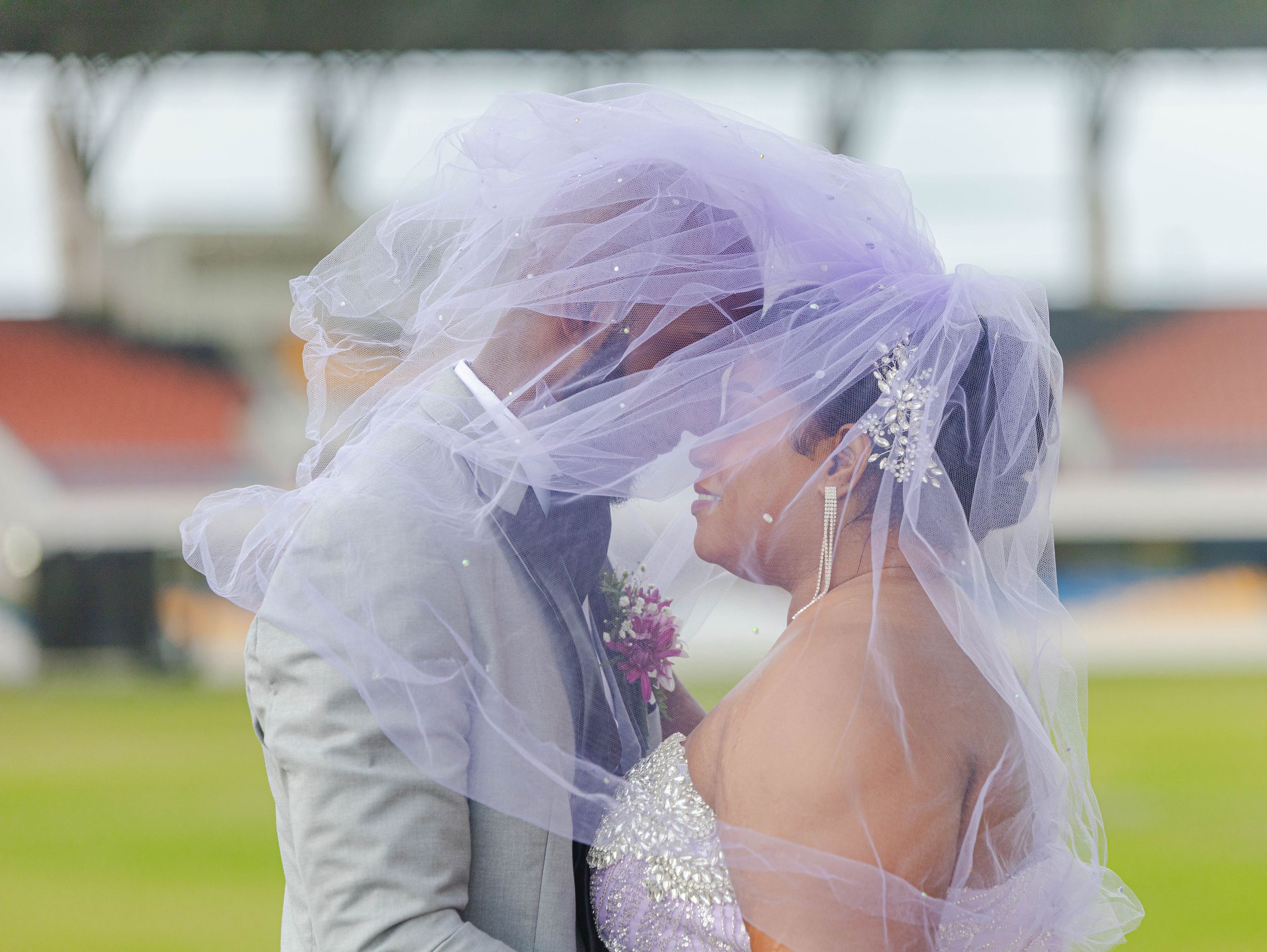 A bride and groom share a romantic kiss beneath a flowing lavender wedding veil outdoors.