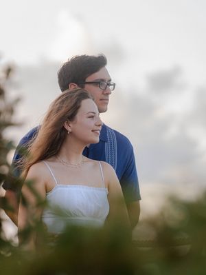 A young couple stands together outdoors, smiling softly, with a blurred green and sky background.