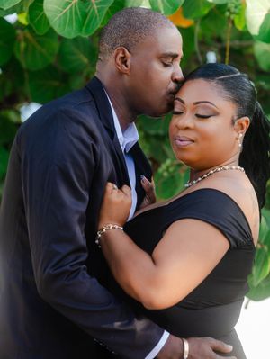 A couple shares a romantic kiss outdoors, the man in a suit and woman in a black off-shoulder dress, with lush greenery behind them.
