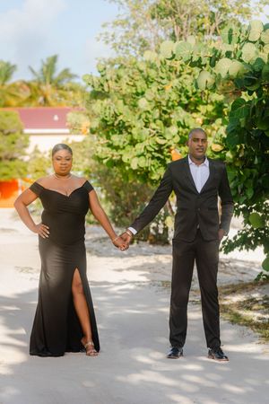 Elegant couple in matching black formal wear holding hands outdoors on a sandy path with tropical greenery.