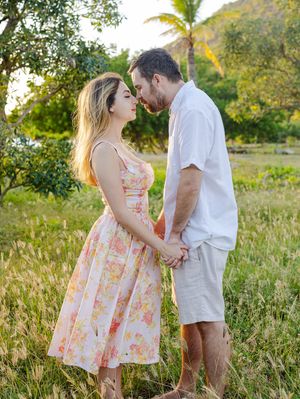 Couple holding hands in a sunny tropical field, woman in floral dress and man in white shirt, sharing a romantic moment.