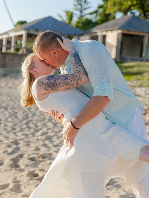 Couple sharing a romantic dip kiss on a sandy beach, man in light blue shirt and woman in white dress.