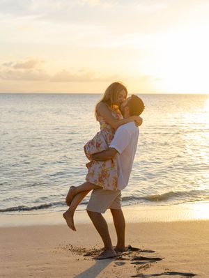 Couple embracing on a sandy beach at golden sunset, man lifting woman in floral dress by the ocean shore.
